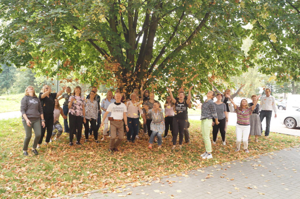 Na zdjęciu grono pedagogiczne pod lipą w pobliżu szkoły. Nauczyciele trzymają liście jesienne lub rzucaja liśćmi i podskakują do góry. Na twarzy mają uśmiech.