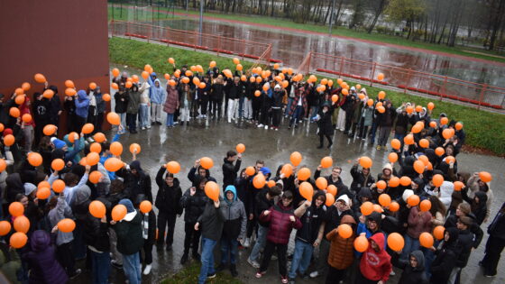 Na zdjęciu młodzież na dziedzińcu szkoły z pomarończowymi balonikami symbolizuje sprzeciw przeciwko przemocy wobec młodziezy