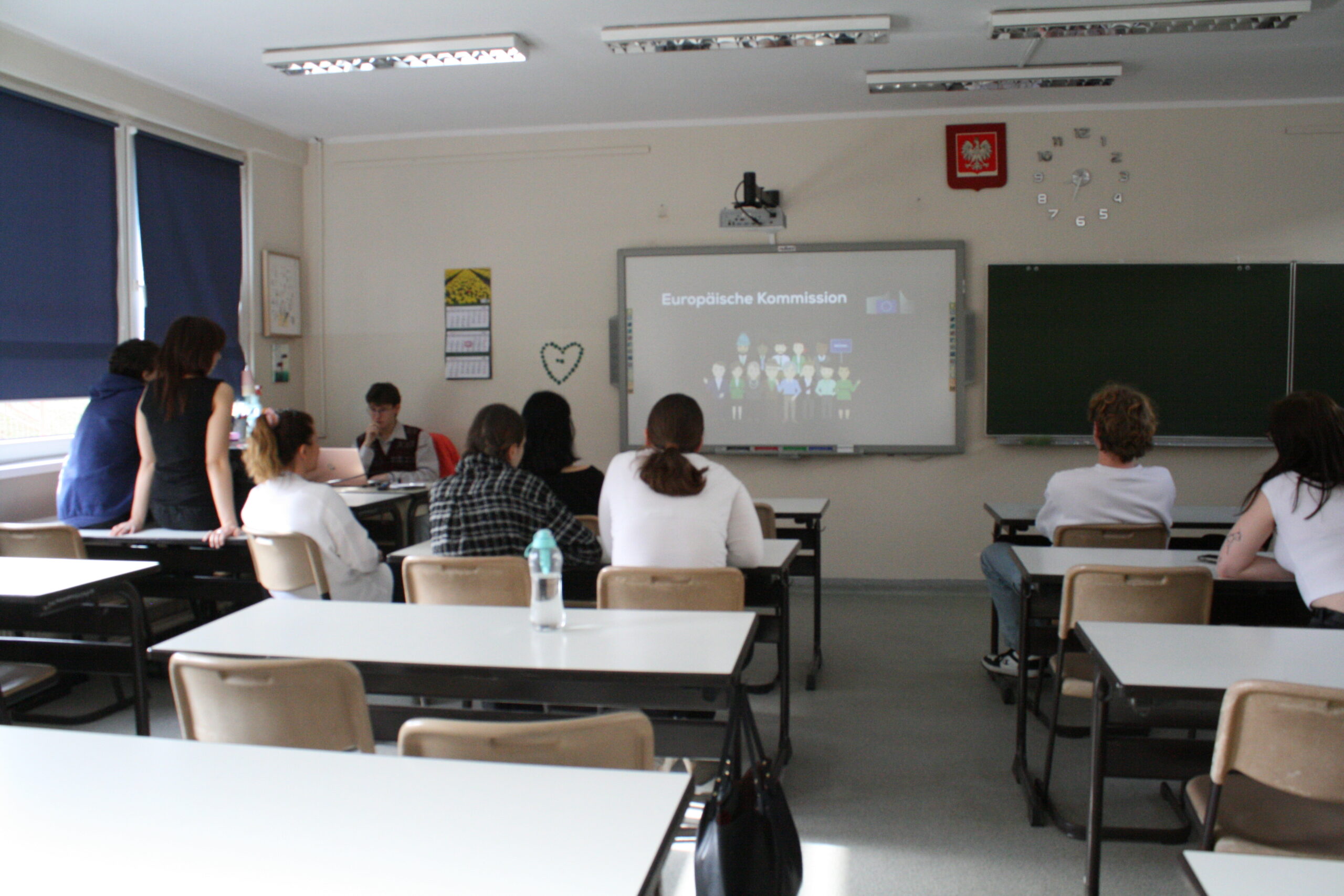 W sali 70 siedzą uczniowie i nauczyciel przed tablicą interaktywną, uczeń siedzi przy biurku