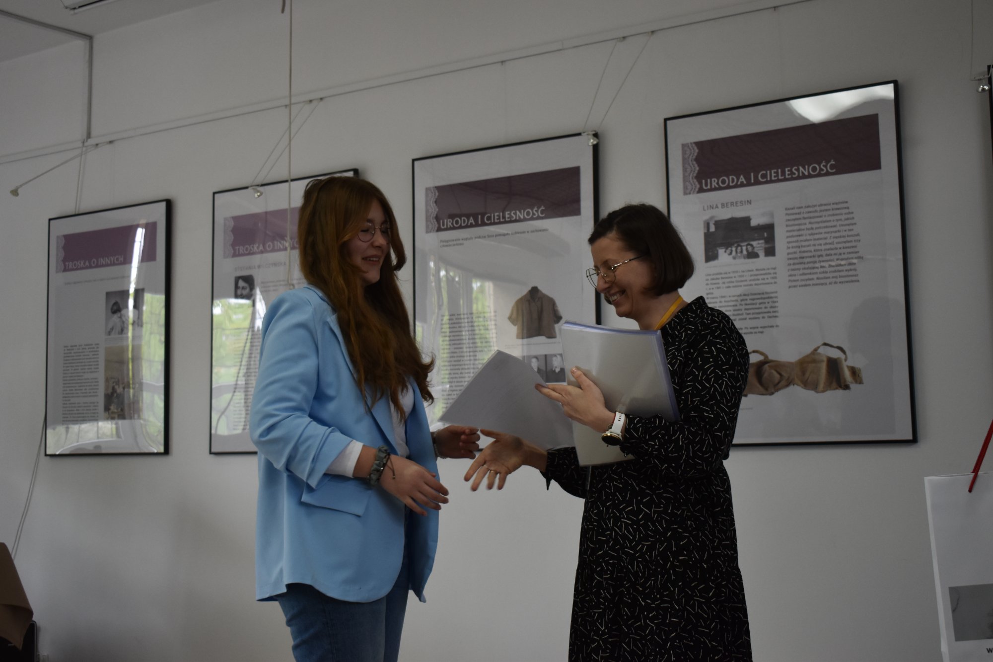 Wolontariuszka otrzymuje dyplom od parcowniczki Państwowego Muzeum Auschwitz -Birkenau, obie na scenie w tle obrazy